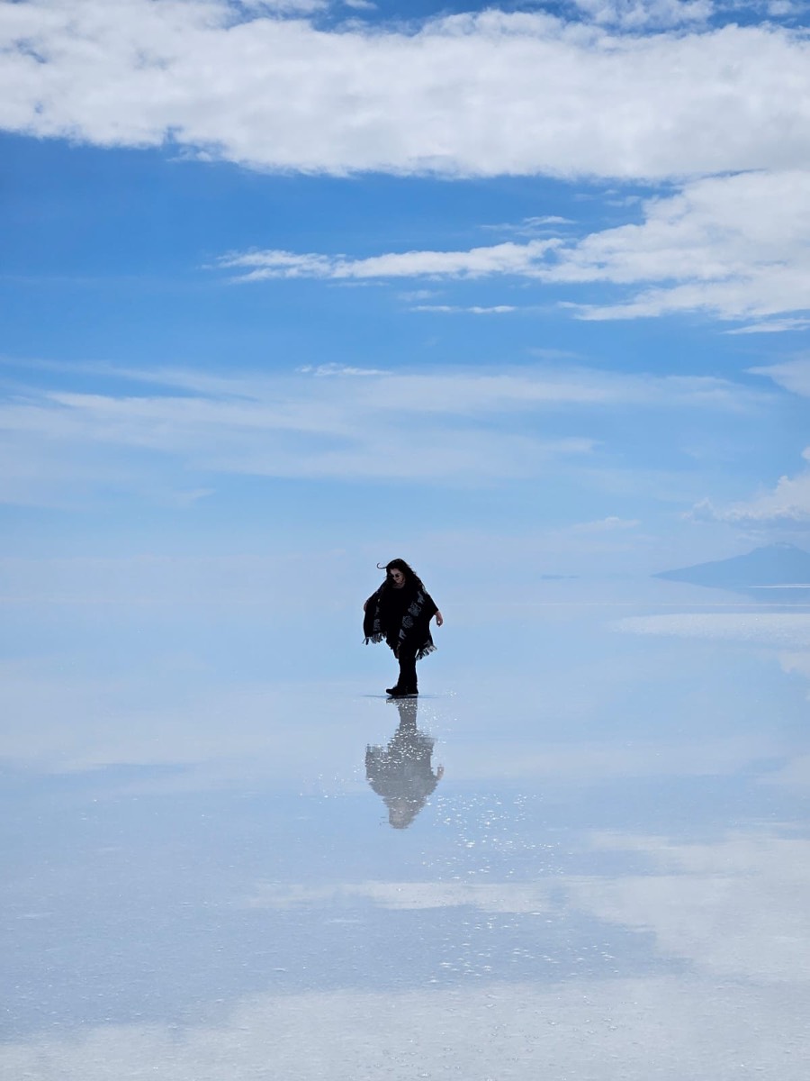 A person walking on a mirrored salt flat under a blue sky with clouds.